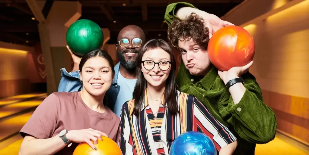 Four friends pose together holding colorful bowling balls at a bowling alley.