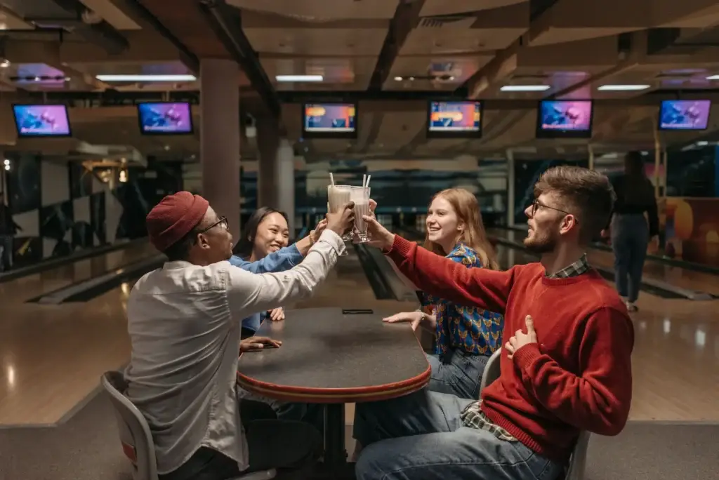 Four friends raise milkshakes in a toast at a table inside a bowling alley.