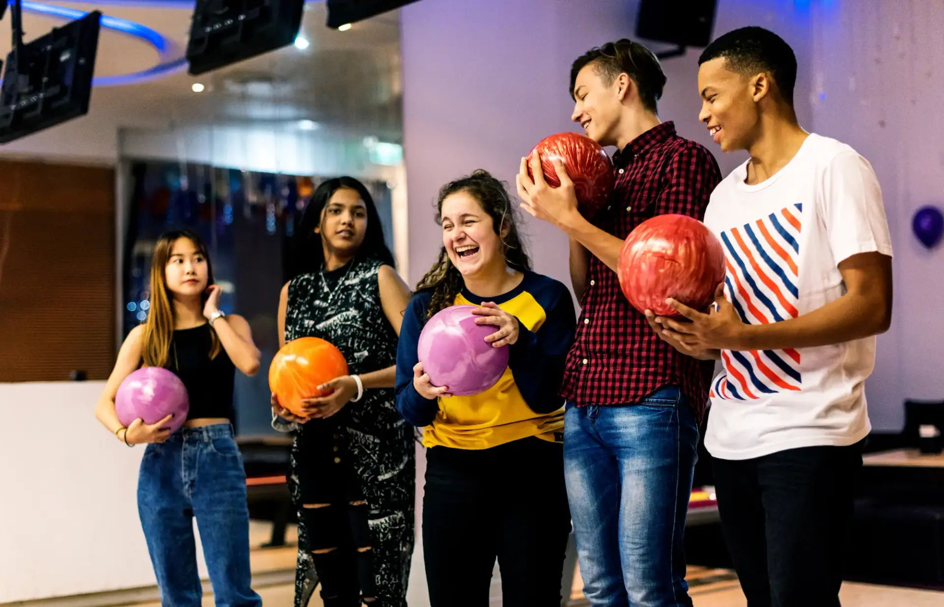 A group of teens laugh together while holding colorful bowling balls.