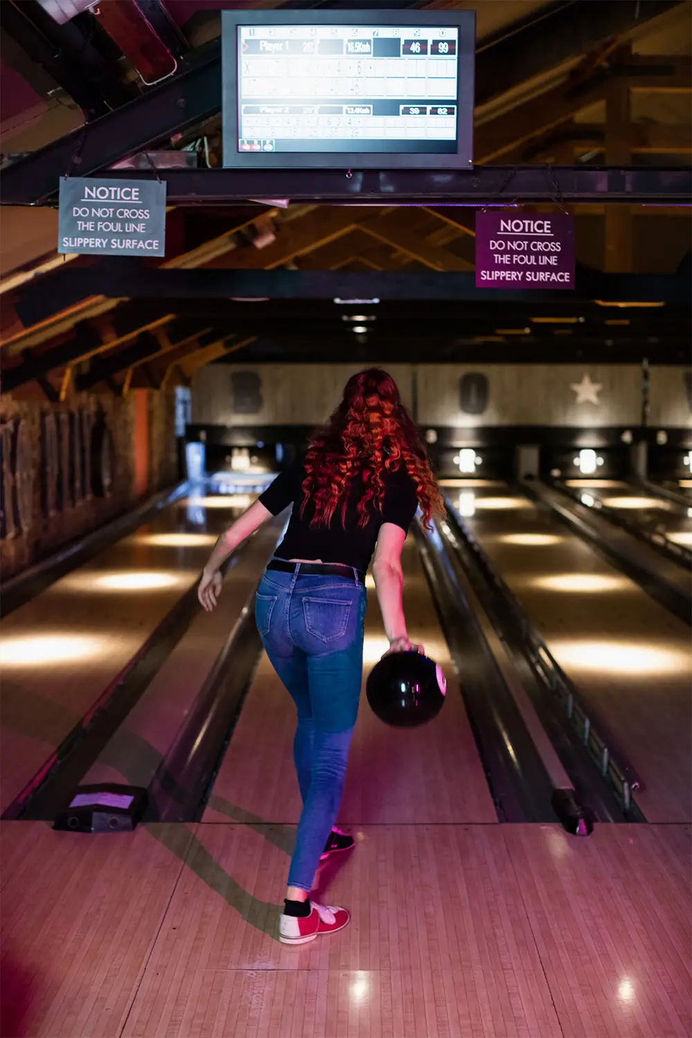 Woman rolling a bowling ball down the lane from behind