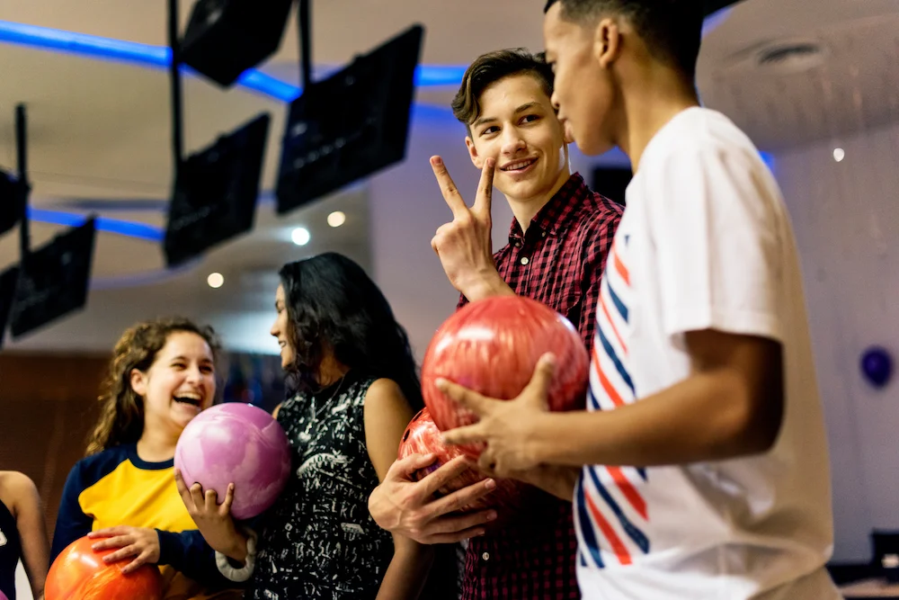Four young friends laughing and holding bowling balls at a bowling alley, one flashing a peace sign.