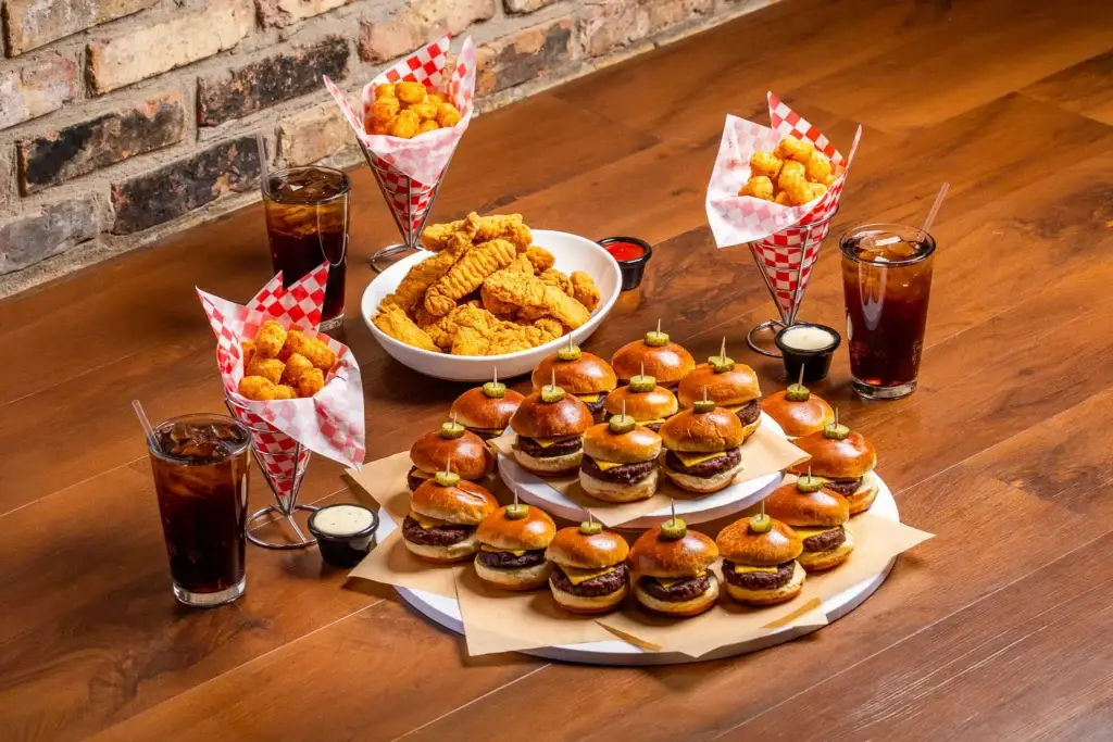 Sliders, chicken tenders, and tater tots arranged on a wooden table with drinks.