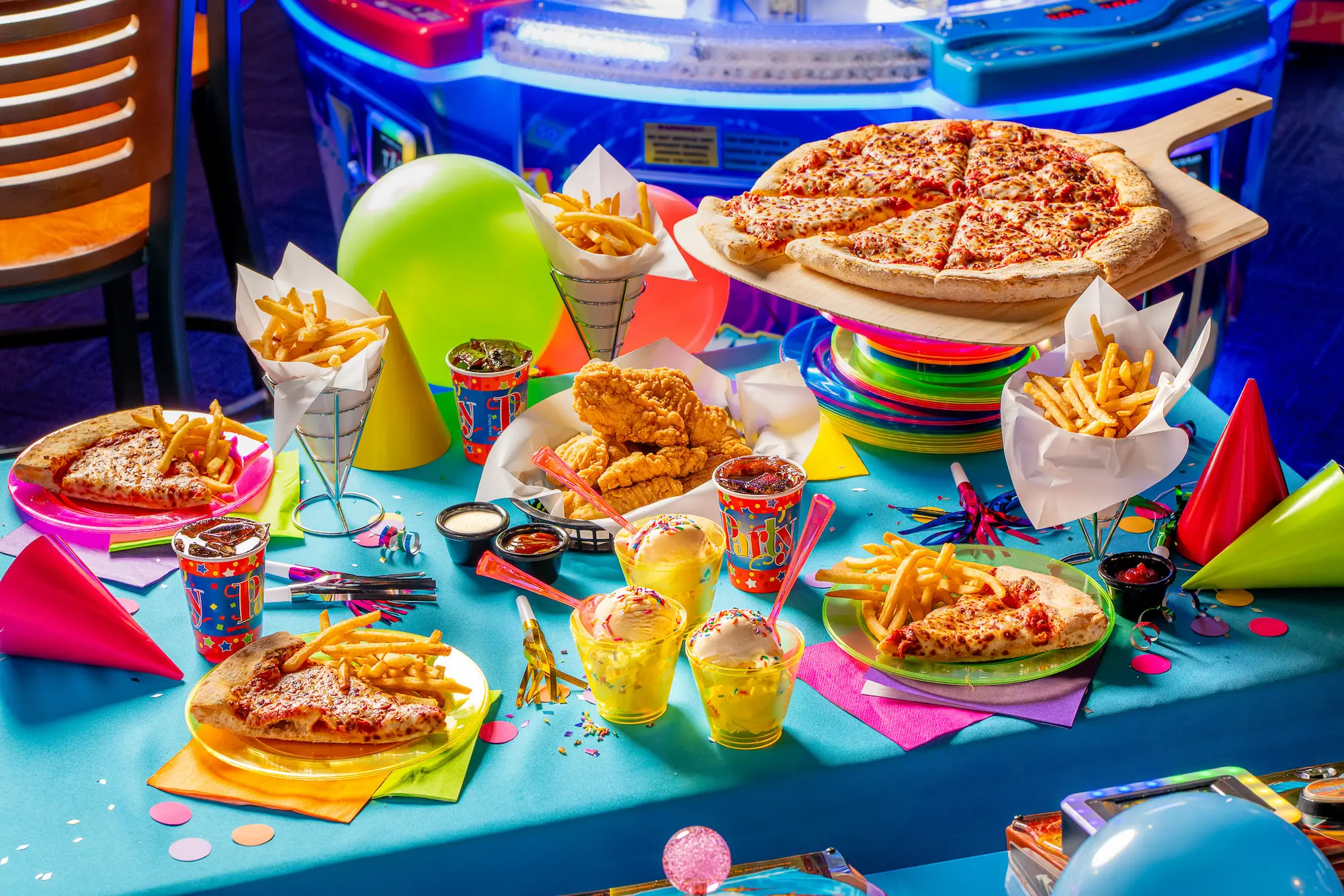 Colorful party food spread including pizza, fries, chicken tenders, and ice cream with balloons and party hats.