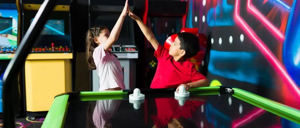 Two kids high-five over an air hockey table at an arcade.