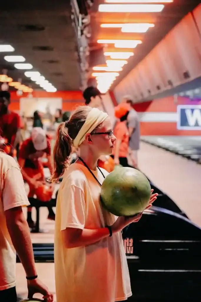 A teenage girl in glasses holds a green bowling ball while waiting at a bowling alley.