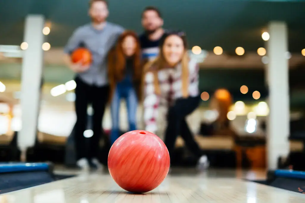 A pink bowling ball sits in focus while a group of friends stand blurred behind it.