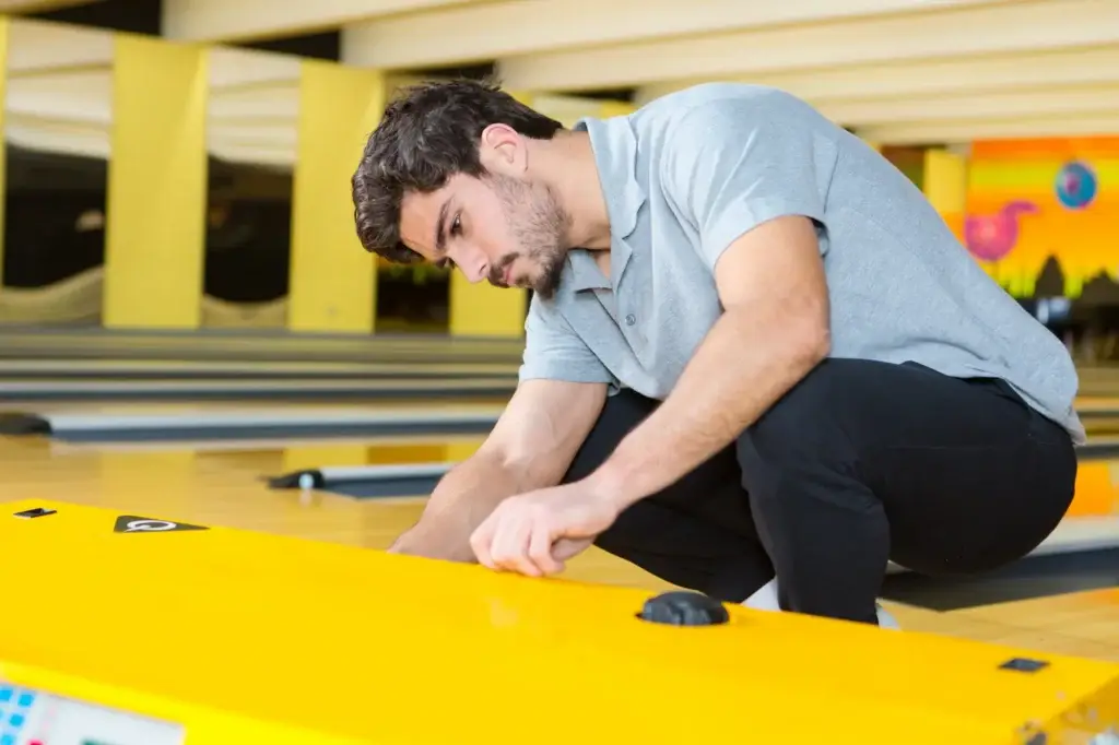 A man crouches and leans on a yellow divider between bowling lanes.