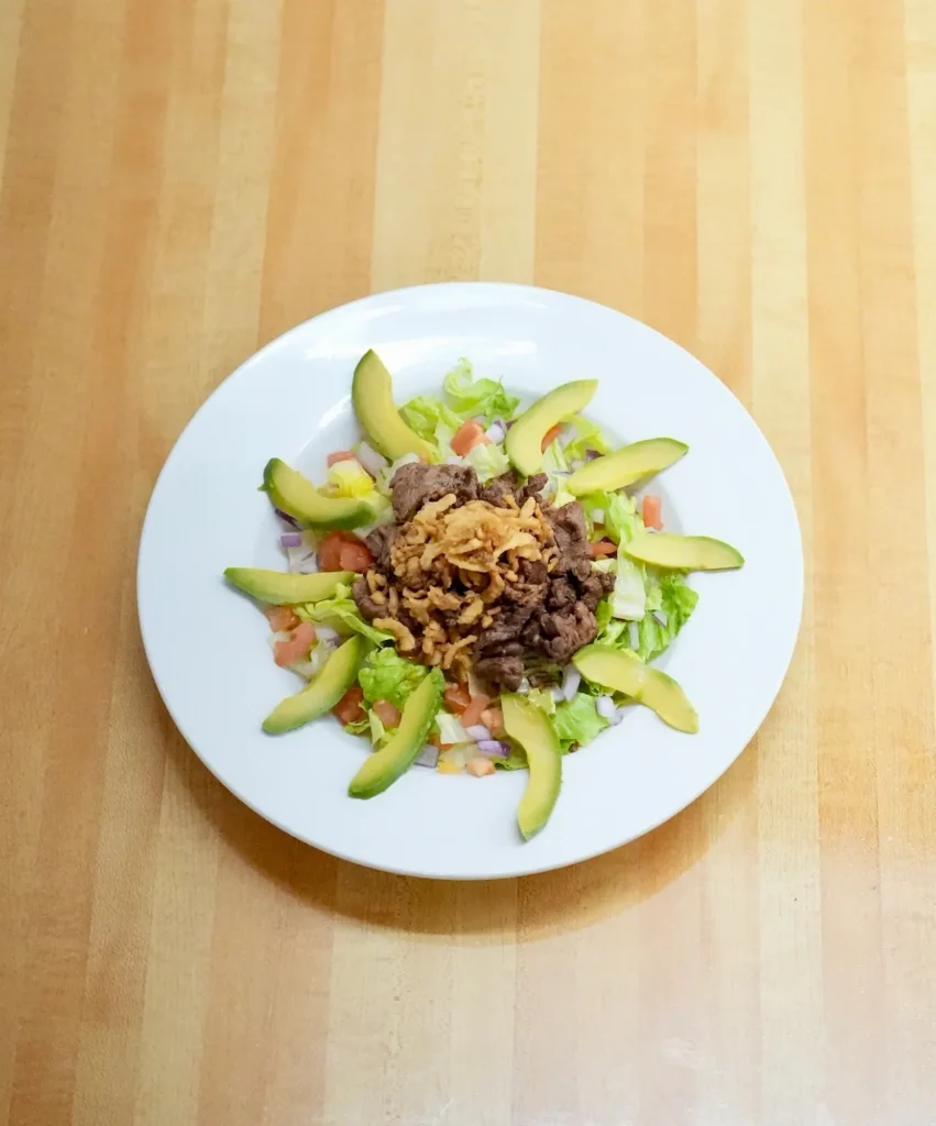Overhead view of Chopped Steak salad topped with avocado slices, steak, tomatoes, red onion, and onion straws on a white plate