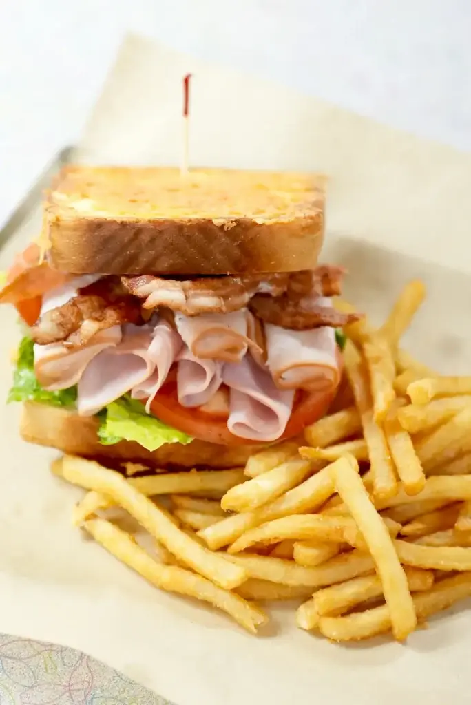 Stacked sandwich filled with sliced pork belly, lettuce, and tomato served with golden crispy fries