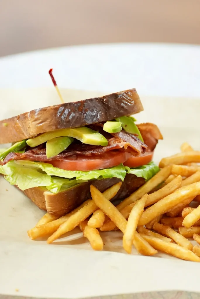 Toasted sandwich loaded with avocado slices, crispy bacon, lettuce, and tomato served with a side of fries