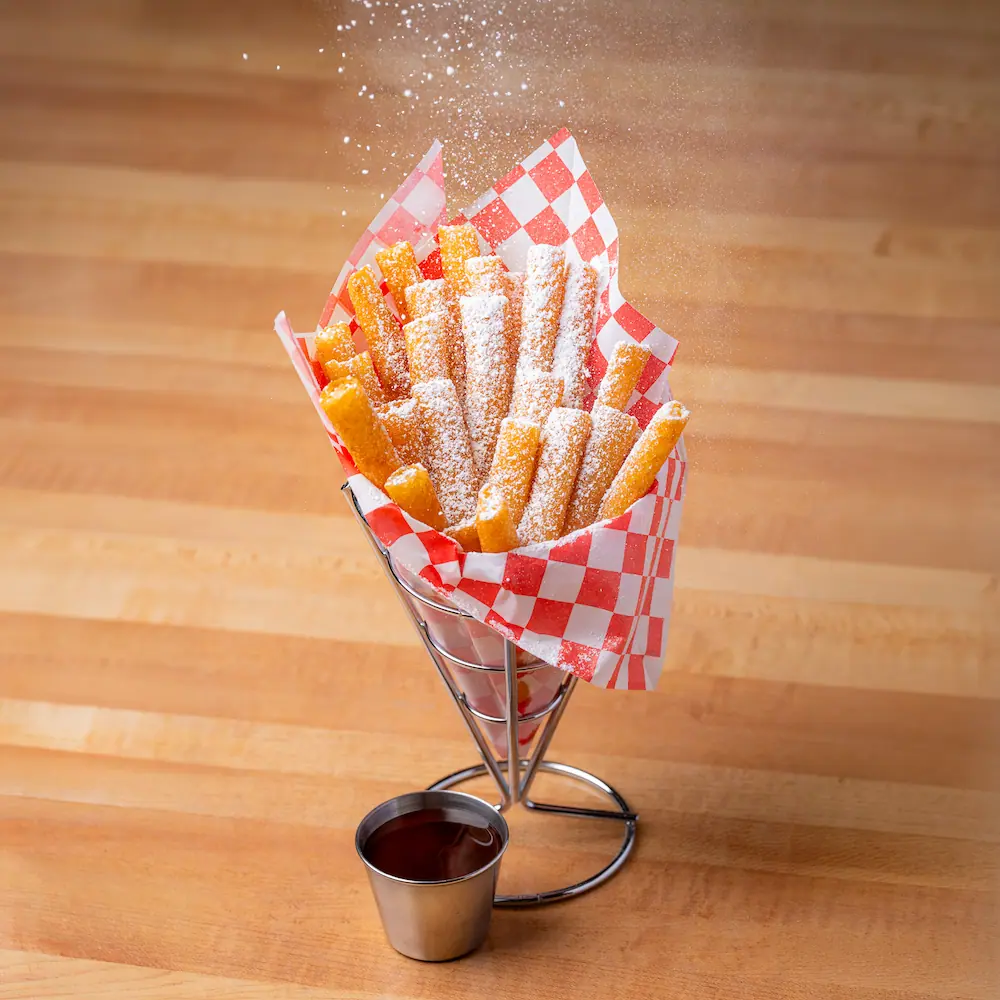 Golden funnel cake fries coated in powdered sugar served in a cone-shaped red checkered liner on a wooden surface
