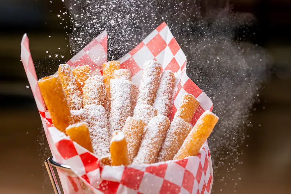 Crispy funnel cake fries dusted with powdered sugar served in a red checkered lined basket with chocolate dipping sauce