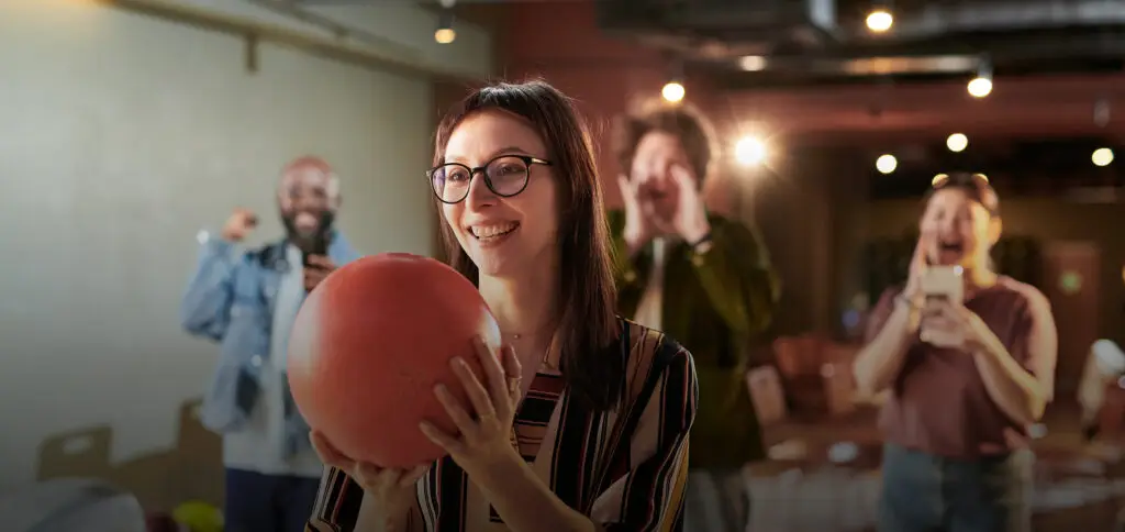 A woman wearing glasses smiles while holding an orange bowling ball.